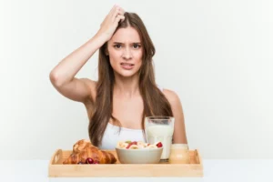 Sieviete apjukusi pie brokastu galda — piena glāze, pārslas un maize; izteikts apjukums par uzturu, ja trūkst olbaltumvielas.