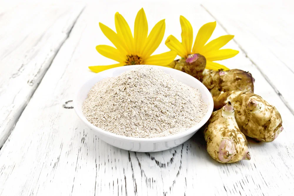 Topinambūru pulveris baltā bļodā ar svaigiem topinambūriem un ziediem uz balta galda
